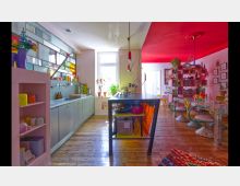 Offene Wohnküche mit einem farbenfrohen, modernen Design. Die Küche verfügt über eine Arbeitsfläche mit Hängelampen, hellgraue Schränke, ein großes Fenster und bunte Regale; der Essbereich ist mit gemusterten Möbeln, Pflanzen und kräftigen rosenroten Akzenten gestaltet.