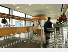 Eingangsbereich mit Drehkreuz und Holzverkleidung, ergänzt durch Metallgeländer. Im Hintergrund sind große Fenster, ein Notausgangsschild und dekorative Blumengestecke sichtbar.