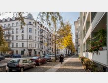 Straßenszene in einem städtischen Wohngebiet mit historischen Altbauten auf der linken Seite und modernen Gebäuden mit Balkonen auf der rechten Seite. Der Gehweg ist von Autos gesäumt, und herbstlich gefärbte Bäume säumen die Allee.