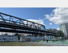Eine Eisenbahnbrücke aus Stahl mit Graffiti-bedeckten Trägern überspannt einen urbanen Raum, der teilweise mit Gras, Gehwegen und einer Skatepark-ähnlichen Fläche ausgestattet ist. Im Hintergrund sind moderne Wohngebäude und Wolken am blauen Himmel zu sehen.