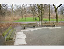 Mehrere Holzbänke stehen auf einem gepflasterten Platz in einem Park. Im Hintergrund befinden sich Laubbäume ohne Blätter, eine Wiese und ein Fußweg, der von einem Metallzaun begrenzt wird.