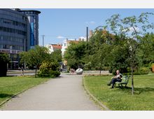 Eine städtische Parkanlage mit Rasenflächen, Bäumen und einem befestigten Weg. Im Hintergrund sind moderne und historische Gebäude sowie eine Werbefläche auf einem Bürogebäude sichtbar, während eine Person auf einer Bank sitzt und Passanten vorbeigehen.