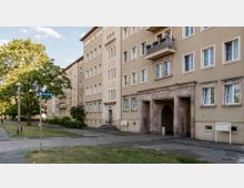 Wohnhaus im Stil der Nachkriegsmoderne mit sandfarbener Fassade, großen rechteckigen Fenstern und einem Torbogen als Eingangsbereich. Davor eine Grünfläche mit einem Baum, Gehwegen und einer Straßenlaterne, Straßenschild "Brunnenring" sichtbar.