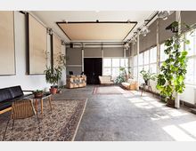 Ein großer, heller Raum mit industriellem Charakter, gekennzeichnet durch Betonboden, hohe Fenster und weiße Wände. Der Raum ist möbliert mit einem Ledersofa, Sesseln, Holztischen und Teppichen, während zahlreiche Topfpflanzen für eine grüne Akzentuierung sorgen.