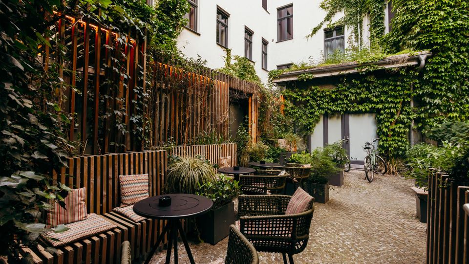 Innenhof mit kleinem Cafébereich, gepflastertem Boden und mehreren runden Tischen mit geflochtenen Stühlen und Sitzkissen. Die hellen Gebäudefassaden und Holzelemente sind stark mit Efeu und Pflanzen bewachsen; am Rand stehen Pflanzkübel und ein Fahrrad.