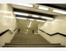 Treppenaufgang in einer U-Bahn-Station mit weißen gefliesten Wänden, schwarzem Handlauf und Deckenleuchten, die den Bereich gleichmäßig beleuchten.