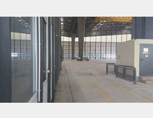 Große industrielle Lagerhalle mit hohen Decken, sichtbaren Stahlträgern und großen Fensterflächen. Ein Teil des Innenraums zeigt eine abgetrennte Wand mit einer Tür und Geländern, während die Halle überwiegend leer ist.