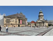 Weitläufiger Platz mit historischem Konzerthaus auf der linken Seite, dessen Dach von einem Baugerüst umhüllt ist, und dem Französischen Dom mit seiner markanten Kuppel rechts im Bild. Menschen spazieren über den gepflasterten Platz bei sonnigem Wetter.