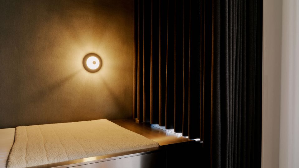 Minimalistischer Innenraum mit einer gepolsterten Liegefläche auf einem Metallpodest, beleuchtet von einer runden Wandleuchte an einer dunkel getönten Wand. Rechts hängen schwere, bodenlange Vorhänge als Raumteiler neben einer hellen Seitenwand.