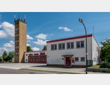 Feuerwehrgebäude mit einem weißen Hauptgebäude und roten Fensterrahmen, daneben ein hoher, rechteckiger Übungsturm aus gelbem Backstein. Vor dem Gebäude befinden sich mehrere rote Garagentore für Einsatzfahrzeuge sowie eine gepflasterte Einfahrt.