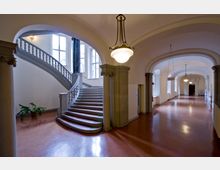 Großzügiger Innenraum eines historischen Gebäudes mit einem geschwungenen Treppenaufgang aus Stein, flankiert von verzierten Säulen. Der Boden ist aus rötlichem Material, und ein langer Korridor mit Gewölbedecken und klassischen Hängelampen erstreckt sich nach rechts.