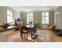 Helles Esszimmer mit Holzboden und grünen Wänden, ausgestattet mit einem großen Holztisch, mehreren Stühlen, einer Kommode und einem Korbsessel. Drei große Fenster lassen Tageslicht herein, während eine Schale mit Obst, Blumen in einer Vase und Deko-Objekte auf dem Tisch und der Kommode den Raum schmücken.