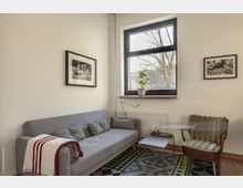 Wohnzimmer mit minimalistischer Einrichtung, bestehend aus einem grauen Sofa mit Kissen, einem Beistelltisch, einem Sessel und einem großen Fenster, durch das Bäume draußen sichtbar sind. An der Wand hängen zwei gerahmte Schwarzweiß-Fotografien, und auf der Fensterbank steht eine kleine Zimmerpflanze.