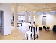 Offener, moderner Innenraum mit Holzboden, einzelnen Säulen und großen Fenstern mit Rundbögen. Der Raum ist mit Tischen, Stühlen, Hockern und Sesseln in einem minimalistischen Stil möbliert, während die helle Beleuchtung und die hohen Decken eine luftige Atmosphäre schaffen.