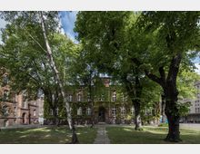 Backsteingebäude mit großen Fenstern, teilweise von Efeu bewachsen, umgeben von hohen Bäumen in einem grünen Park; im Hintergrund sind weitere Gebäude sichtbar.