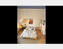 Schlafzimmer mit einem Holzbett, ungemachtem Bettzeug und hellem Parkettboden. An der Wand hängen Bilder, ein großer Spiegel lehnt an der rechten Seite, und eine Papierlampe hängt von der Decke.