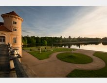 Schlosspark mit einer beigefarbenen Schlossfassade mit Rundturm im Vordergrund; symmetrisch angelegter Garten mit geschwungenen Wegen, Rasenflächen und Statuen, im Hintergrund ein ruhiger See mit Baumreihe entlang des Ufers bei Sonnenuntergang.