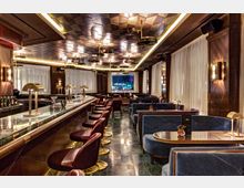 Luxuriöse Bar mit einer langen, marmorgekleideten Theke, umgeben von gepolsterten roten Barhockern mit goldenen Fußstützen. Der Raum ist geprägt von dunklen Holzwänden, weißen Vorhängen, einem kunstvoll gestalteten Deckendesign und gemütlichen Sitzbereichen mit blauen Polstern und goldfarbenen Akzenten.