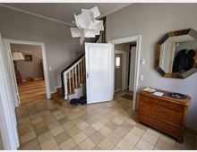 Heller Eingangsflur eines Wohnhauses mit großformatigen beigefarbenen Bodenfliesen, grauen Wänden und einer Holztreppe mit weißem Geländer. Mehrere offene Türen führen in angrenzende Räume; an der Wand hängen ein achteckiger Spiegel, daneben steht eine Holzkommode, und an der Decke hängt eine skulpturale weiße Pendelleuchte.