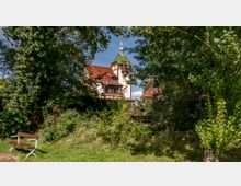Ein historisches Gebäude mit einem grünen Turmdach und roten Ziegeldächern ist von Bäumen und dichter Vegetation umgeben. Im Vordergrund steht eine Holzbank auf einer Wiese.