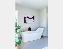 Modernes Badezimmer mit einer freistehenden weißen Badewanne in geschwungener Form vor einer hellen Wand. Ein dekoratives Kunstbild hängt an der Wand, und links im Bild steht eine große grüne Zimmerpflanze auf dem Boden.