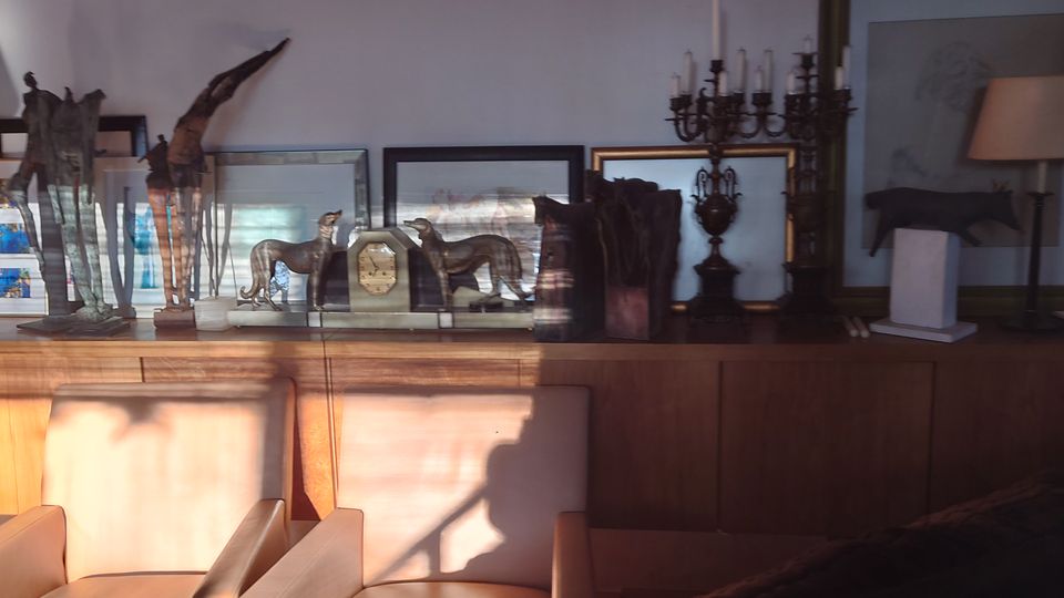 Ein Wohnzimmer mit Holzmöbeln, darunter ein Sideboard mit dekorativen Objekten wie Skulpturen, Kerzenständern und gerahmten Bildern. Im Vordergrund stehen gepolsterte Stühle, und warmes Licht fällt durch ein Fenster in den Raum.