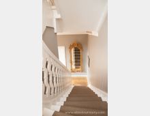 Treppe in einem hellen Innenraum mit weißem Geländer und beigefarbenem Teppichläufer; am unteren Ende der Treppe ein großer, goldener Spiegel mit verziertem Rahmen, der einen Teil des Raumes reflektiert. Die Wände sind in einem neutralen Grauton gestrichen.