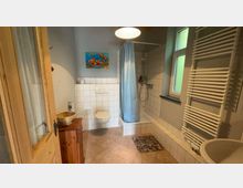 Ein schlichtes Badezimmer mit beiger Bodenfliese und weißen Wandfliesen, ausgestattet mit einer Duschkabine mit blauem Vorhang, einem WC, einem Waschtisch, einem großen weißen Handtuchheizkörper und einem Fenster. Ein buntes Bild mit einem Fischmotiv hängt über dem WC, und ein Holzschränkchen steht links im Raum.