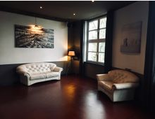 Wohnraum mit minimalistischem Design, ausgestattet mit zwei weißen Polstersofas auf einem rotbraunen Boden. Große Fenster lassen natürliches Licht herein, und Wände sind mit gerahmten Schwarz-Weiß-Bildern dekoriert; eine Stehlampe steht in der Ecke.