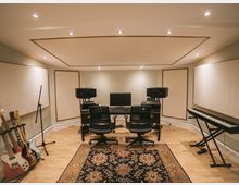 Ein professionelles Musikstudio mit zwei Stühlen und einem zentralen Tisch, auf dem ein Computer und Lautsprecher stehen. Links steht ein Gitarrenständer mit mehreren E-Gitarren und ein Mikrofonständer, rechts ein elektronisches Keyboard; der Raum ist mit schallabsorbierenden Paneelen und einem großen Teppich gestaltet.