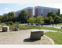 Platz mit gepflasterter Fläche und großen Steinen im Vordergrund, im Hintergrund mehrere Wohnblöcke mit farbigen Fassaden in Weiß, Orange und Rot, umgeben von Bäumen und einer ruhig verlaufenden Straße.