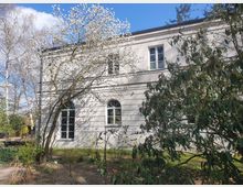 Heller Gebäudeflügel mit weißen Wänden und großen, gewölbten Fenstern, umgeben von blühenden Bäumen und Sträuchern in einem Garten. Im Hintergrund sind kahle Bäume und ein blauer Himmel zu sehen.