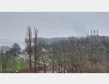 Blick auf eine winterliche Landschaft mit kahlen Bäumen, einem Sportplatz im Vordergrund und einem See im Hintergrund. Rechts am Horizont steht ein Industriegebäude mit drei Schornsteinen.