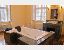 Luxuriöses Badezimmer mit einer großen, rechteckigen Badewanne in einer Verkleidung aus grauem Marmor. Im Hintergrund befinden sich ein Waschtisch mit Spiegel, zwei große Fenster mit weißen Rahmen und Heizungselemente.