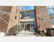 Backsteinfassade eines mehrstöckigen Gebäudes mit großen, verglasten Fenstern und vergitterten Türen im Erdgeschoss. Im Vordergrund liegen Holzstapel und diverse Gegenstände, umgeben von einem Betonboden und weißem Gestänge.