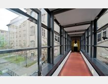 Verbindungsflur mit gläsernen Wänden und schwarzem Metallrahmen, der zwischen zwei Gebäuden verläuft. Im Hintergrund ist ein historisches Gebäude mit hellen Fassaden und zahlreichen Fenstern sichtbar, umgeben von einem begrünten Innenhof mit blühenden Bäumen.