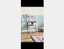 Helles Zimmer mit einer blau gestrichenen Wand, einem kleinen weißen Schreibtisch und einem weißen Bürostuhl mit Fellüberzug. An der Wand befinden sich zwei schwebende Regale mit Dekorationsgegenständen; der Boden ist aus Holz im Fischgrätmuster, und ein orientalischer Teppich bedeckt teilweise den Boden.