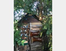 Ein kleines, handgefertigtes Holzhaus steht zwischen dichten Bäumen. Die Fassade ist aus unregelmäßigem Holz gefertigt und teilweise mit dekorativen Elementen wie Glasbausteinen und Rindenstücken verziert.