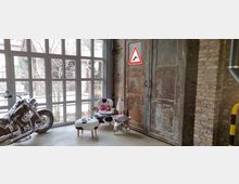 Ein industriell gestalteter Innenraum mit großen Fensterfronten, durch die ein Garten sichtbar ist. Links steht ein Motorrad, rechts eine alte Metalltür vor einer Ziegelwand, daneben dekorative Möbel mit Kuhfellbezug und ein Warnschild mit Hirschsymbol.