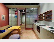 Modern gestaltete Küchen- und Essraum-Kombination mit offenem Design; links eine Sitzbank mit gelbem Tisch und bunten Hockern, rechts eine minimalistische Küchenzeile mit Regalen und Holzarbeitsplatte. Hintergrund mit Glasbausteinen und Holztrennwänden, die einen weiteren Raum andeuten.