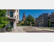 Straßenszene mit einer Kreuzung, links ein Altbau mit abgerundetem Balkon und rechts ein modernes Gebäude mit grauer Fassadenverkleidung. Im Hintergrund steht eine große rote Backsteinkirche mit einem spitzen Turm, umgeben von Bäumen und einer Grünfläche.