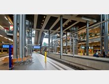 Moderne Bahnhofsplattform mit breitem Bahnsteig, hohen Decken und großen Glas- und Metallkonstruktionen. Mehrere Ebenen mit Rolltreppen, Aufzügen und Geschäften im Hintergrund; Anzeigetafeln und Sitzbänke sind entlang des Bahnsteigs sichtbar.