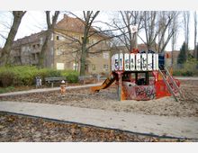Spielplatz in einer herbstlichen Umgebung mit einem Holzspielgerüst in Form eines kleinen Bootes, das mit Graffiti bemalt ist. Im Hintergrund sind Wohngebäude mit roten Ziegeldächern und gelb umrahmten Fenstern sowie Bäume ohne Laub erkennbar.