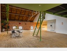 Loftartiger Innenraum mit Backsteinwänden, Holzbalkendecke und großen, bogenförmigen Fenstern. Zentraler Essbereich mit einem Tisch und Drahtstühlen, grüne Metallleiter zu einer oberen Ebene, sowie eine offene, moderne Küchennische im Hintergrund.