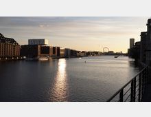 Flusslandschaft bei Sonnenuntergang mit modernisierter Uferbebauung, darunter Backstein- und Neubauten, auf beiden Seiten des Wassers; im Hintergrund rauchende Schornsteine und ein Industriebereich. Ein Geländer ist im rechten Vordergrund sichtbar.