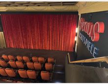 Innenraum eines kleinen Kinos mit roten Samtvorhängen vor der Leinwand, orangefarbenen Polstersitzen in Reihen und einem Treppengeländer im Vordergrund. An der rechten Wand hängt ein beleuchtetes Schild mit der Aufschrift „Venus Lichtspiele“.