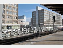 Ansicht eines Bahnhofs mit Bahnsteigen und Gleisen im Vordergrund. Im Hintergrund befinden sich moderne, mehrstöckige Gebäude mit rechteckigen Fassaden und großen Fenstern, sowie Geschäfte und Schilder entlang der Straße.