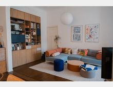 Bright, modern living room with a built-in wooden shelving wall filled with books and decor, a pale blue sectional sofa with colorful cushions, and a low round coffee table on a light area rug. White walls feature two abstract framed artworks, with a paper globe pendant light overhead and a small plant near a closed door.