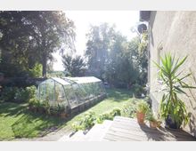 Ein Garten mit einer Rasenfläche, einem Glashaus voller Pflanzen und mehreren Blumentöpfen im Vordergrund. Rechts steht ein beigees Wohngebäude mit einer großen Yucca-Palme in einem Topf auf einer hölzernen Terrasse, und im Hintergrund sind Bäume und Sträucher zu sehen.