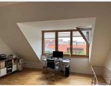 Dachgeschossbüro mit einem Schreibtisch vor einem großen Fenster mit Blick auf rote Ziegeldächer und Bäume. Der Raum hat schräge Wände, einen Holzfußboden, ein Regal mit Ordnern auf der linken Seite und eine Wäschetrockner-Leine rechts.